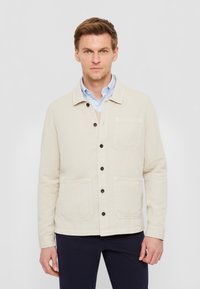 Giacca in tela beige con colletto, dotata di bottoni neri, due tasche frontali e un taglio dritto. Indossata sopra una camicia azzurro chiaro.