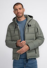 Chaqueta acolchada en verde oliva, con capucha, bolsillos con cremallera negros y una textura ligeramente brillante. Lleva encima de una sudadera azul.