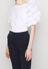 Valkoinen t-paita, jossa on teksturoitu ja röyhelöity hiha toisella puolella. Yhdistetty tummansinisiin suoralahkeisiin housuihin, joissa on suora leikkaus ja vyö.