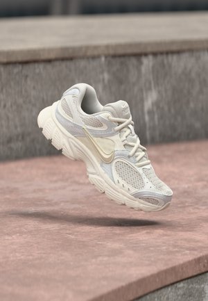 Zapatilla de correr Nike blanca y beige con paneles de malla y suela gruesa, suspendida sobre una superficie de piedra rosa y un fondo de piedra gris.