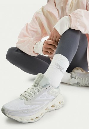 Osoba sedící v tureckém sedu, obutá v světle šedých teniskách Reebok, bílých ponožkách Reebok, tmavých legínách a světle růžové bundě.