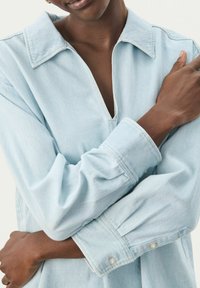 Camicia di jeans blu chiaro con colletto a corolla, scollatura a V profonda e maniche lunghe. Presenta polsini con bottoni e una vestibilità rilassata con cuciture visibili.