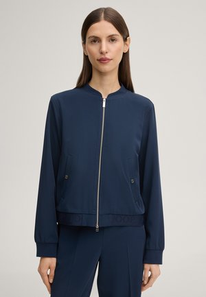 Donna che indossa una giacca blu navy con zip, tasche laterali con bottoni e pantaloni blu navy abbinati, in piedi davanti a uno sfondo chiaro e uniforme.