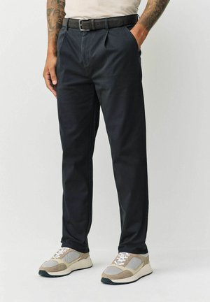 Black pleated trousers with a straight cut, paired with a black belt. Worn with beige and gray sneakers featuring textured accents.