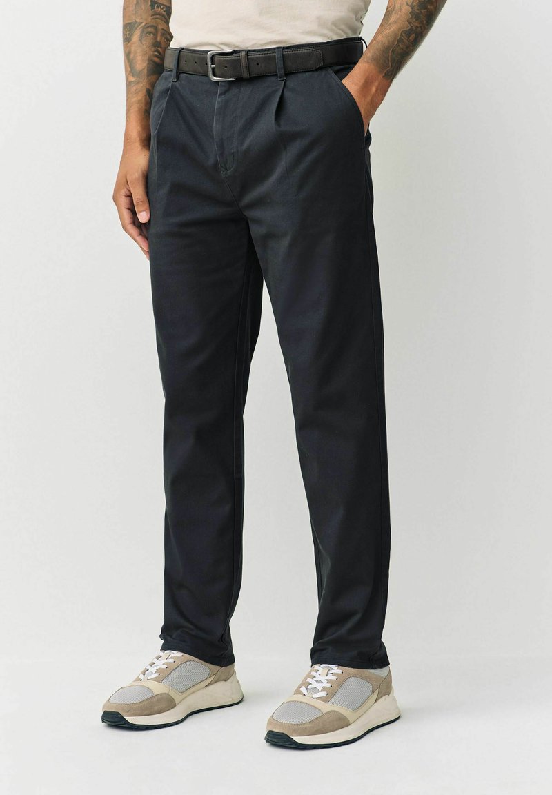 Black pleated trousers with a straight cut, paired with a black belt. Worn with beige and gray sneakers featuring textured accents.