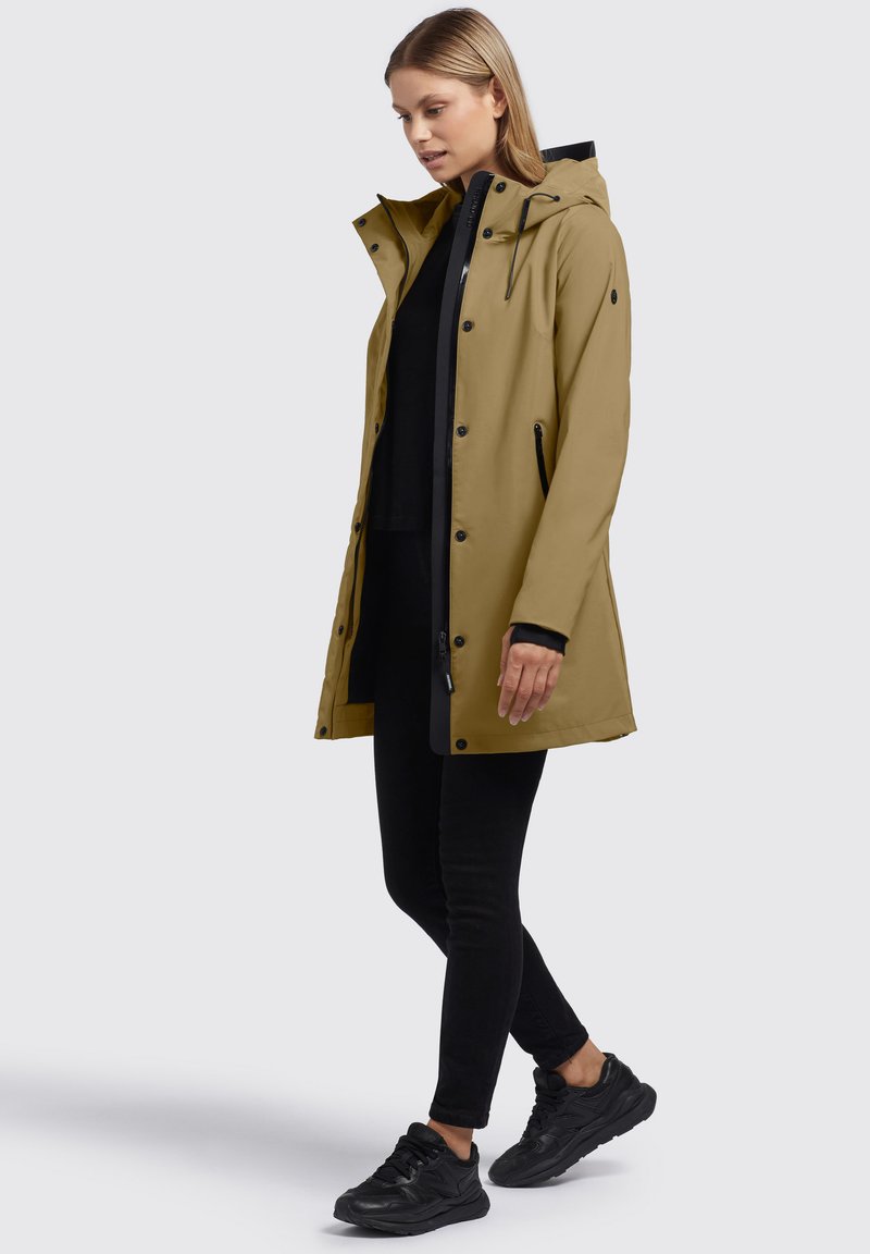 Veste imperméable kaki avec capuche, boutons-pression à l'avant et accents noirs, associée à un pantalon slim noir et des baskets noires. Texture lisse.