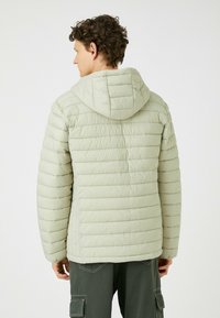 Lys grøn pufjakke med horisontal quiltning, en hætte og elastiske manchetter. Lavet af glat, vandafvisende materiale.