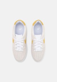 Vita mockask sneakers med gula accenter, med perforerad tå, vita snören och en vadderad sula med Nike-logotyp.