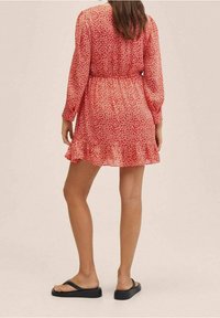 Rotes Blumenkleid mit langen Ärmeln und gerüschtem Saum, aus leichtem Stoff gefertigt. Getragen mit schwarzen Flip-Flops und tailliertem Schnitt.
