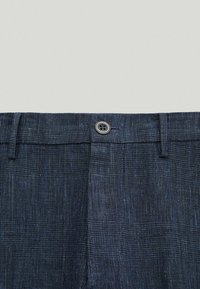 Granatowe spodnie z teksturowanym, kratkowanym wzorem. Posiadają zapięcie na guzik oraz szlufki na pasek. Materiał ma subtelny połysk.