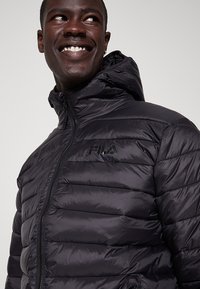 Giacca a vento nera con zip frontale e cappuccio. Presenta una texture trapuntata e il logo "FILA" sul lato sinistro del petto. Materiale liscio e lucido.