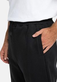 Schwarze Sweatpants aus weichem Material, mit elastischem Bund, Seitentaschen und lockerer Passform. Hand ruht zur Detailbetonung in der Tasche.