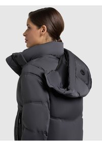 Tmavě šedá péřová bunda s velkou skládací kapucí. Má zipové kapsy a prošívanou strukturu, která poskytuje teplo a ochranu.