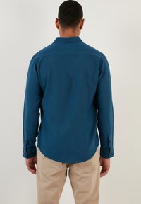 Camisa de manga larga azul oscura con cuello abotonado, corte recto y puños de un solo botón, vista desde la parte posterior. Textura de tela suave.