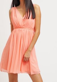 Šaty bez rukávů meruňkové barvy s výstřihem do V, nařaseným pasem a splývavou, plisovanou sukní. Vyrobené z lehkého, hladkého materiálu.