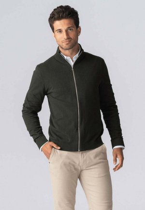 Mørkegrøn zip-up sweater med en ståkrave, ribbede manchetter og kant. Båret over en hvid skjorte med brune bukser. Blød tekstur.