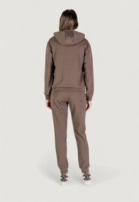 Mujer con cabello castaño largo, vistiendo una sudadera marrón y pantalones a juego, zapatillas blancas con tacones de patrón oscuro, de pie mirando hacia atrás.