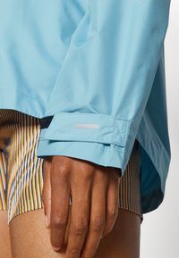 Manica di giacca impermeabile azzurro chiaro con chiusura in velcro nera, dotata di un accento riflettente e pantaloni corti a strisce in contrasto sotto.