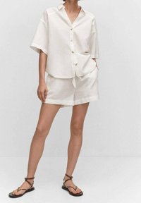 Chemise beige clair à manches courtes, boutonnée à l'avant, portée avec un short assorti. Portée avec des sandales noires à lanières.