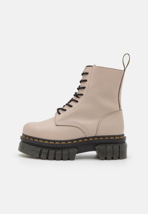 Botte de combat beige à lacets avec semelle noire épaisse à rainures, couture jaune et languette de tirage au talon, vue de profil sur fond blanc.