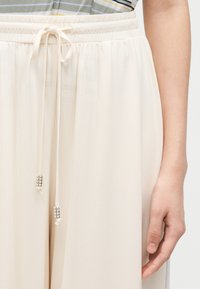 Κοντινή λήψη ενός ατόμου που φοράει κρεμ παντελόνι με κορδόνι στη μέση με χάντρες στις άκρες και ριγέ μπλούζα, δείχνοντας τη μέση και το δεξί χέρι.