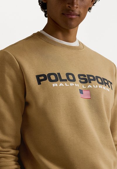 Khaki krēklveida džemperis ar apaļu kakla izgriezumu, kurā ir redzams uzraksts "POLO SPORT" un "RALPH LAUREN" treknā melnā burtiem, kā arī neliels Amerikas karoga akcents.