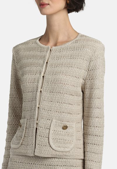 Femeie purtând un cardigan bej din tricot cu model zigzag texturat, cu guler rotund, nasturi în față și două buzunare cu clapetă și nasturi aurii.