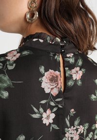 Floresal blouse sisältää mustaa kangasta, jossa on vaaleanpunaisia ja vihreitä kukkakuvioita, rypytetyn kaulan, sekä avaimenreikän muotoisen selän mustilla nappikiinnityksillä.