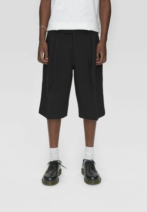 Zwarte geplooide shorts met een losse fit, met een strak ontwerp en rechte zoom. Draag ze met witte sokken en zwarte veterschoenen.