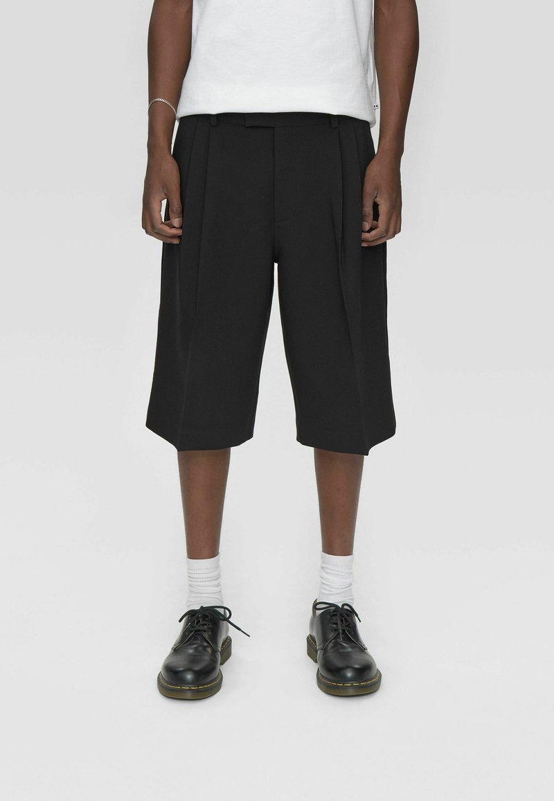 Shorts plissés noirs avec une coupe ample, présentant un design épuré et un ourlet droit. Portés avec des chaussettes blanches et des chaussures noires à lacets.