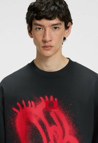 Czarna bluza z czerwonym nadrukiem w plamy i czerwonym logo. Posiada okrągły dekolt oraz detal z ściągaczami przy rękawach.