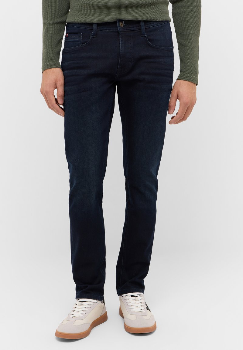 Ciemnoniebieskie dżinsy o slim fit, z pięcioma kieszeniami, zapinane na guzik, z delikatnym przetarciem, w połączeniu z jasnymi tenisówkami.