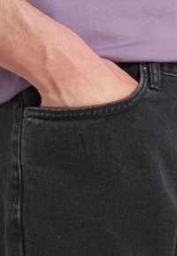 Jeans de mezclilla negra con una vista en primer plano que muestra una mano en el bolsillo. El material tiene una ligera textura y un diseño clásico de cinco bolsillos.