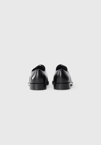 Une paire de chaussures de costume en cuir noir poli, vues de derrière, sur un fond gris clair uni.