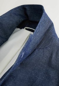 Nærbillede af en mørkeblå denimjakke med synlige syninger, hvidt indvendigt for og en knaphul på reversen.