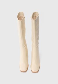 Des bottes en cuir crème montantes, à bouts carrés et avec des zips sur le côté, présentées debout sur un fond blanc.