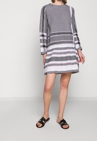 Vestido de rayas y diseños geométricos variados en blanco y negro, con mangas largas y cuello redondo. Se lleva con sandalias negras.