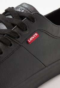 Fekete szintetikus sportcipő texturált felülettel, fekete fűzőkkel és piros Levi's címkével az oldalon. Alacsony szabású, gumitalppal.