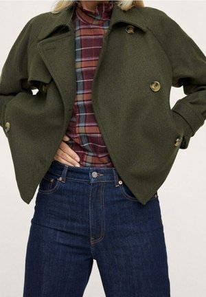 Kobieta ubrana w oliwkowo zielony, krótki płaszcz na czerwoną i czarną koszulę w kratę, zestawioną z wysokim stanem ciemnoniebieskich dżinsów.