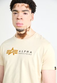 Beidzis kokvilnas T-krekls ar tumšu logotipu, kurā rakstīts "ALPHA INDUSTRIES" ar zelta akcentiem, īsām piedurknēm un apaļu dekoltē.