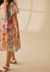 Abito a fantasia floreale in colori pastello, con maniche corte arricciate, design a strati e orlo ondulato; indossato con sandali bianchi.