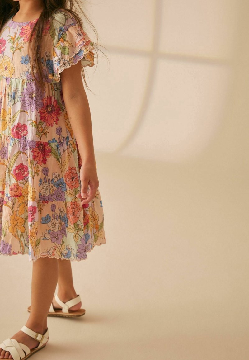 Abito a fantasia floreale in colori pastello, con maniche corte arricciate, design a strati e orlo ondulato; indossato con sandali bianchi.