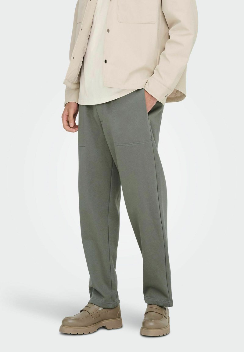 Veste beige clair par-dessus une chemise crème, associée à un pantalon ample vert olive avec deux poches avant, sur un fond neutre.