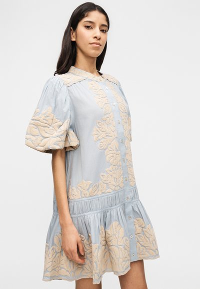Νεαρή γυναίκα που φοράει ένα ανοιχτό γαλάζιο φόρεμα με κουμπιά, διακοσμημένο με μπεζ ανθικό κέντημα και φουσκωτά κοντά μανίκια, στέκεται μπροστά σε ένα απλό φόντο.