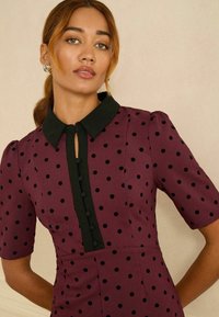 Burgundivörös ruha fekete pöttyökkel, rövid ujjal, fekete gallérral és kulcslyuk gombzárral. Simább anyag, formázott illeszkedéssel.