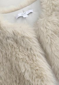 Tissu en fausse fourrure beige doux avec une étiquette de vêtement blanche portant la mention "NEXT" cousue dans une doublure intérieure de couleur claire.