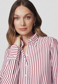 Kobieta z jasnobrązowymi falującymi włosami nosi złote geometryczne kolczyki oraz białą koszulę z guzikami w pionowe czerwone paski, opierając rękę na szyi.