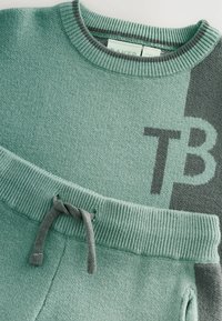 Žalias megztinis su kontrastinėmis pilkomis detalėmis, su grioveliais apatinėje dalyje ir reguliuojamu raišteliu. Išskirtinė logotipo detalė.