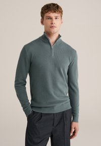 Žalias megztinis su užtrauktuku ant kaklo, tekstūruotu paviršiumi, ilgomis rankovėmis ir gofruotomis rankogaliais. Derinamas su tamsiomis kelnėmis, suteikiant pilną įvaizdį.