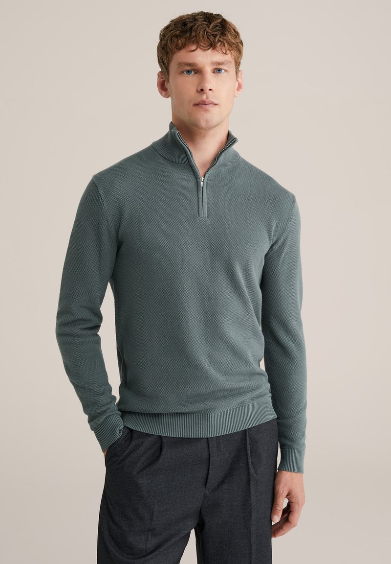 Žalias megztinis su užtrauktuku ant kaklo, tekstūruotu paviršiumi, ilgomis rankovėmis ir gofruotomis rankogaliais. Derinamas su tamsiomis kelnėmis, suteikiant pilną įvaizdį.